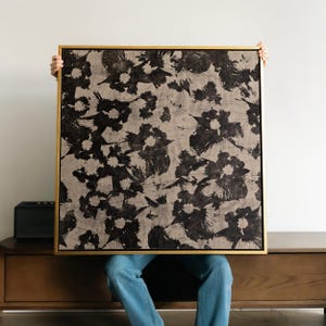 Op de afbeelding: Een ingelijst kunstwerk met een herhalend patroon van zwarte bloemvormen op een beige achtergrond. Het kunstwerk wordt door een persoon omhoog gehouden, waarbij de gouden lijst te zien is. Het stuk heeft een moderne, abstracte uitstraling.