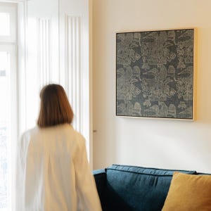 May include: A framed wall art piece with a dark blue background and a repeating pattern of birds and floral designs in a light beige color. The artwork is displayed on a white wall above a blue sofa with a yellow pillow.