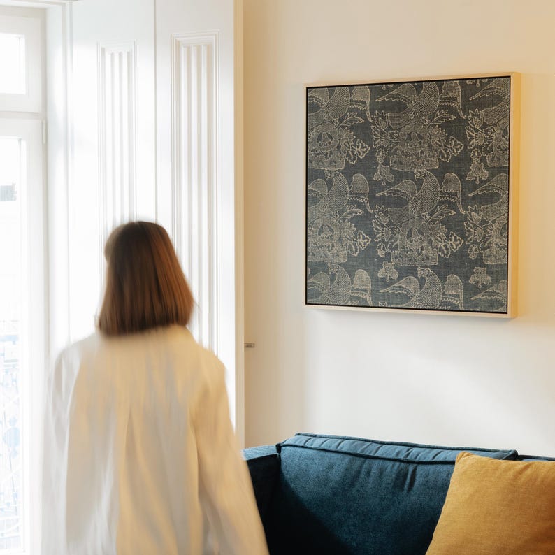 May include: A framed wall art piece with a dark blue background and a repeating pattern of birds and floral designs in a light beige color. The artwork is displayed on a white wall above a blue sofa with a yellow pillow.
