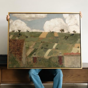 May include: A framed landscape painting in a gold-coloured frame. The artwork shows a rural scene with rolling green hills, patches of yellow fields, and buildings with red roofs under a cloudy sky. The painting is held by a person wearing blue jeans.