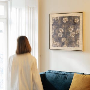 May include: A framed wall art print featuring a dark blue background with a pattern of white and gold floral designs. The artwork is displayed in a black frame and hangs on a white wall above a blue sofa.