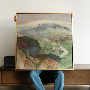 May include: A square framed wall art piece featuring an abstract landscape painting with textured layers of muted green, brown, blue, and beige tones. The artwork is enclosed in a thin, metallic gold frame, showcasing a soft, hazy horizon line.