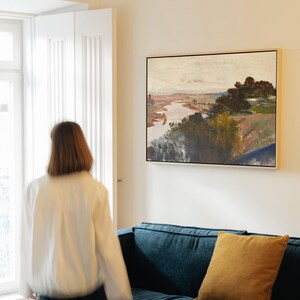 May include: A framed landscape painting depicting a river and trees. The artwork uses a muted colour palette with shades of brown, green, and beige. The painting is above a blue sofa with a yellow cushion.
