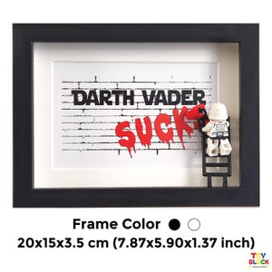 Può includere: Una scatola ombra incorniciata in nero con una stampa di un muro di mattoni bianchi che presenta il testo "Darth Vader Sucks" in vernice rossa. Una minifigure Lego di uno Stormtrooper sta dipingendo il testo su una scala.