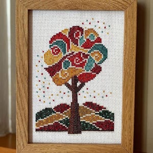 Puede incluir: Obra de arte de punto de cruz de un árbol con un diseño abstracto y colorido. La copa del árbol presenta remolinos rojos, amarillos y turquesas, con un tronco y una base marrones. La obra está enmarcada en un marco de madera marrón claro.