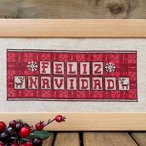 May include: Cross-stitch artwork in a wooden frame. The design features the words "FELIZ NAVIDAD" in white block letters on a red plaid background. Snowflakes and reindeer flank the text. A sprig of red berries rests in the lower left corner.