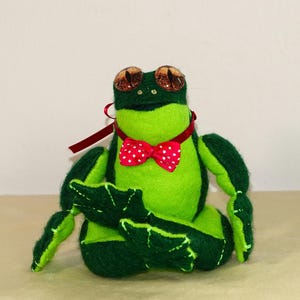 May include: A handmade plush frog in shades of green, with large, textured eyes and a red bow tie with white polka dots. The frog has a red ribbon around its neck and is sitting with its legs outstretched.