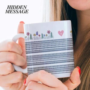 Puede incluir: Taza de cerámica blanca con un diseño floral y geométrico. La taza presenta un corazón rosa con una cruz y el texto "HIDDEN MESSAGE". El diseño incluye flores coloridas y patrones en blanco y negro.