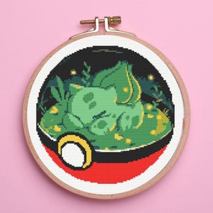 May include: A circular wooden embroidery hoop containing a cross-stitch pattern of a green creature sleeping inside a red, white, and black spherical container. The design features pixel art style elements with green foliage and small yellow light accents.
