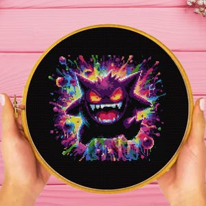May include: A circular wooden embroidery hoop containing a cross-stitch design of a purple, spiky-eared creature with glowing red eyes and a wide, toothy grin, set against a black background surrounded by a vibrant, multi-colored paint splatter effect.