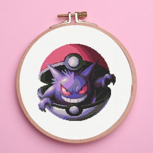 Puede incluir: Un aro de bordado de punto de cruz con un personaje Pokémon morado y negro, Gengar, dentro de una Poké Ball roja y blanca.