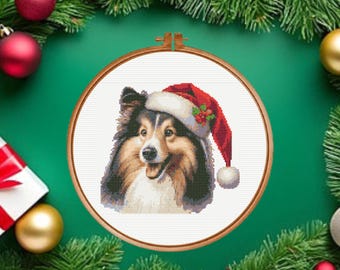 May include: A cross-stitch pattern featuring a Shetland Sheepdog with black, tan, and white fur wearing a red and white Santa hat with a small green holly sprig. The embroidery is displayed within a circular wooden hoop against a green background.