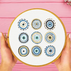 May include: A circular wooden embroidery hoop containing white fabric with a cross-stitch pattern of nine distinct circular motifs. Each motif features concentric rings in shades of blue, tan, brown, and black, arranged in a three-by-three grid.