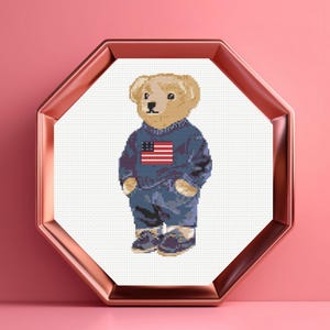 May include: Cross-stitch design of a teddy bear wearing a blue sweatshirt with an American flag and blue pants. The bear is standing inside a white octagon-shaped frame with a rose gold border, set against a pink background.