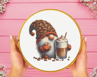 May include: A cross-stitch pattern in a wooden embroidery hoop featuring a gnome wearing a hat covered in coffee bean patterns. The gnome holds a chocolate chip cookie in one hand and a glass of iced coffee with whipped cream in the other.