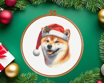 May include: A cross-stitch embroidery of a Shiba Inu dog wearing a red and white Santa hat with a small sprig of holly. The embroidery is contained within a circular wooden hoop, featuring a white background and detailed stitching of the dog.