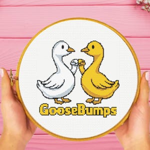 May include: A circular wooden embroidery hoop containing a cross-stitch pattern of a white goose and a yellow goose performing a fist bump. Below the geese, the text GooseBumps is stitched in yellow block letters against a white fabric background.