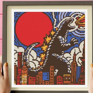 May include: A framed cross-stitch pattern featuring a large dark creature with yellow dorsal fins standing in a city with skyscrapers. A giant red circle sits in the blue sky behind the creature, which is breathing fire toward the right side.