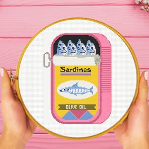 May include: A cross-stitch design of a pink sardine can with the word "Sardines" in yellow. The can contains blue fish and the words "Olive Oil". The design is in a wooden embroidery hoop.