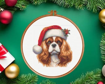 May include: A cross-stitch embroidery pattern featuring a Cavalier King Charles Spaniel wearing a red and white Santa hat with a sprig of holly, displayed within a circular wooden embroidery hoop against a white fabric background.