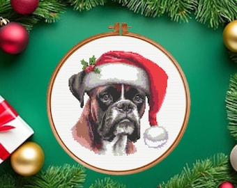 Puede incluir: Un patrón de punto de cruz de un perro bóxer que lleva un gorro de Papá Noel rojo y blanco con una pequeña rama de acebo, exhibido dentro de un bastidor de bordado circular de madera. El perro tiene pelaje marrón y negro con hocico blanco.