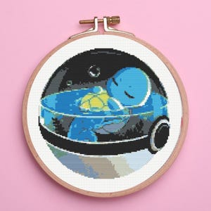 May include: A cross-stitch embroidery project featuring a blue creature with a yellow shell resting inside a spherical container. The design is held within a circular wooden embroidery hoop, showcasing detailed pixel-style stitching on white fabric.