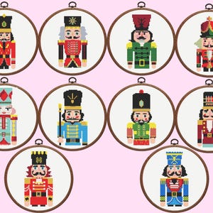 May include: Ten circular wooden embroidery hoops containing cross-stitch patterns of nutcracker figures. Each nutcracker features a unique uniform design in various colours including red, blue, green, and gold, displayed against a white fabric background.