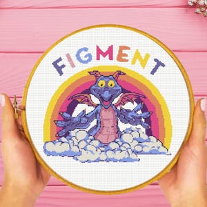 May include: A circular wooden embroidery hoop containing a cross-stitch pattern of a purple dragon character named Figment. The design features the text FIGMENT in colorful letters above a rainbow, clouds, and the dragon figure in a pixel art style.