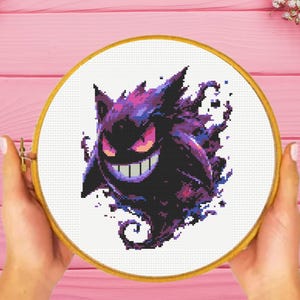 May include: A circular wooden embroidery hoop containing a cross-stitch pattern of a purple and black Gengar character. The design features a pixelated aesthetic with sharp spikes, glowing pink eyes, and a wide, toothy grin, set against a white fabric.