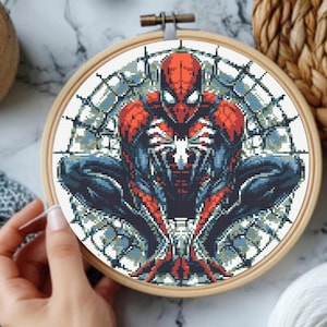 Könnte beinhalten: Ein runder Stickrahmen aus Holz mit einem Kreuzstichmuster eines Superhelden in hockender Pose. Das Design zeigt einen rot-dunkelblauen Anzug mit einem weißen Spinnenemblem auf der Brust, vor einem Hintergrund, der an ein Spinnennetz erinnert.