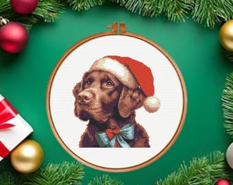May include: A cross-stitch pattern in a round wooden embroidery hoop featuring a brown dog wearing a red and white Santa hat and a light blue bow tie. The design is centered on a white fabric background, showcasing detailed stitching of the dog.