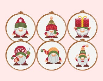 May include: Six circular wooden embroidery hoops containing cross-stitch patterns of gnomes. Each gnome wears a patterned hat in shades of red, green, and yellow, featuring a white beard, and holding various holiday items like gifts and ornaments.