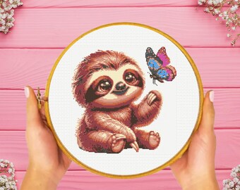 May include: A cross-stitch pattern featuring a brown sloth with large dark eyes sitting and reaching toward a colorful butterfly with blue, pink, and black wings. The design is displayed within a circular wooden embroidery hoop frame.