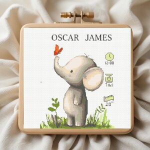 May include: A square wooden embroidery hoop containing a cross-stitch pattern of a light gray elephant with a small orange butterfly on its trunk. The text OSCAR JAMES is at the top, with icons for time, weight in lbs, and length in 20 inches.