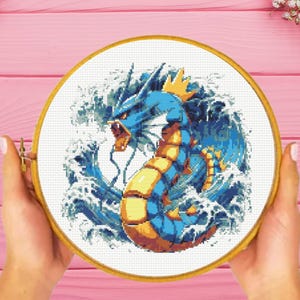 May include: A blue and yellow dragon-like Pokemon character with a crown, surrounded by white waves, is depicted in a cross-stitch pattern.