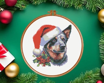 Pode incluir: Um padrão de ponto cruz com um cão boiadeiro-australiano usando um gorro de Pai Natal vermelho e branco, posicionado sobre um grupo de folhas verdes de azevinho e bagas vermelhas, dentro de um bastidor de bordar circular de madeira.