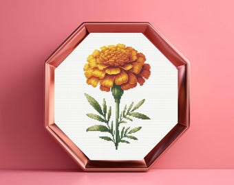 May include: A cross-stitch pattern of a vibrant orange and yellow marigold flower with green leaves and a stem, displayed inside an octagonal metallic copper-coloured frame against a solid pink background.