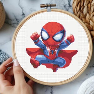 Può includere: Un motivo a punto croce raffigurante un personaggio supereroe in stile chibi pixelato con un costume rosso e blu, grandi occhi bianchi e un mantello rosso fluente, esposto all'interno di un telaio da ricamo circolare in legno.