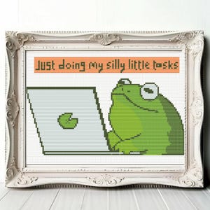 Puede incluir: Un patrón de punto de cruz con una rana verde sentada frente a una computadora con el texto "Just doing my silly little tasks".