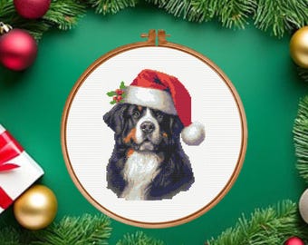 Puede incluir: Un patrón de punto de cruz que muestra un perro de montaña de Berna con un gorro de Papá Noel rojo y blanco con una ramita de acebo. El diseño se presenta en un bastidor de bordado de madera circular sobre un fondo de tela blanca.