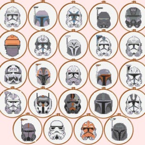 Peut inclure: Un ensemble de 25 motifs de point de croix représentant des casques de soldats clones de Star Wars. Les casques sont de différentes couleurs, notamment blanc, gris, noir et orange. Chaque casque est encadré dans un cercle.