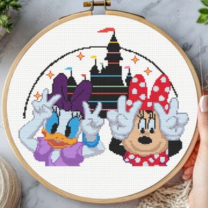 Könnte beinhalten: Ein runder Holzstickrahmen mit einem Kreuzstichmuster von Daisy Duck und Minnie Mouse, die das Friedenszeichen zeigen. Dahinter befindet sich die Silhouette einer schwarzen Burg mit bunten horizontalen Streifen und kleinen Sternen.