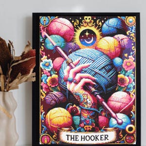 Peut inclure: Une impression d'art murale encadrée représentant une illustration colorée d'une main tenant un crochet et une grosse pelote de laine bleue, entourée de diverses pelotes et fleurs. La bannière inférieure affiche le texte THE HOOKER en noir.