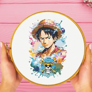 Op de afbeelding: Een kruissteekpatroon met een kleurrijke illustratie van een cartoonfiguur met een strohoed en een schedel en gekruiste beenderen symbool. Het figuur is omringd door kleurrijke verfspatten.