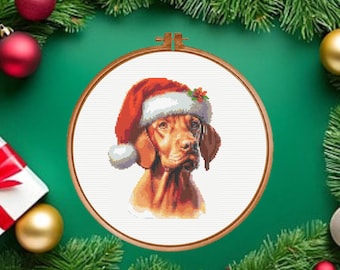 May include: A cross-stitch pattern in a wooden embroidery hoop featuring a brown Vizsla dog wearing a red and white Santa hat with a small green holly sprig. The dog is centered on a white fabric background, showcasing detailed stitching.