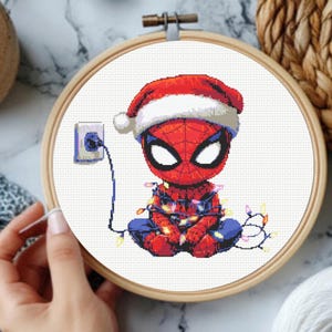 Könnte beinhalten: Ein Kreuzstichmuster in einem hölzernen Stickrahmen, das eine Spider-Man-Figur im Chibi-Stil mit einer rot-weißen Weihnachtsmütze zeigt, die in eine bunte Lichterkette verwickelt ist und an einer Steckdose auf der linken Seite angeschlossen ist.