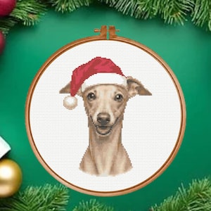 Pode incluir: Um bordado em ponto cruz apresentando um galgo italiano castanho claro usando um gorro de Pai Natal vermelho e branco. O bordado é exibido dentro de um bastidor de madeira circular, mostrando pontos detalhados do rosto do cão.