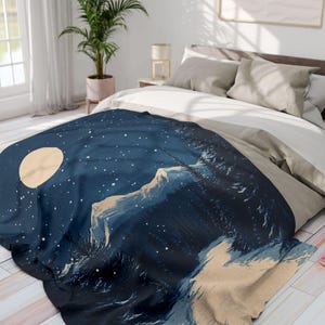 May include: A blue and beige throw blanket featuring a mountain scene and a full moon in the night sky. The blanket has a dark blue background with white stars and a beige mountain range with a forest in the foreground.