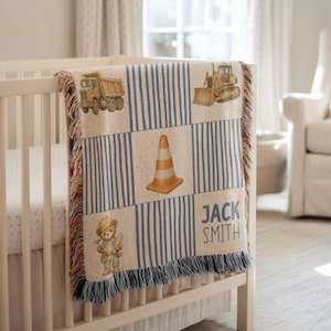Personalized construction-themed woven baby blanket draped over a white nursery crib, featuring watercolor-style construction vehicles including a dump truck, bulldozer, traffic cone, and construction worker teddy bear. The blanket has blue striped patchwork blocks and custom baby name text, creating a soft vintage construction nursery decor aesthetic perfect for baby boys and baby shower gif