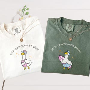 May include: Two t-shirts, one cream and one olive green, with the phrase "off to commit aunt foolery" and "off to commit uncle foolery" printed on them. Each shirt features a cartoon goose wearing a hat and swimsuit. A necklace with a pendant is on each shirt.